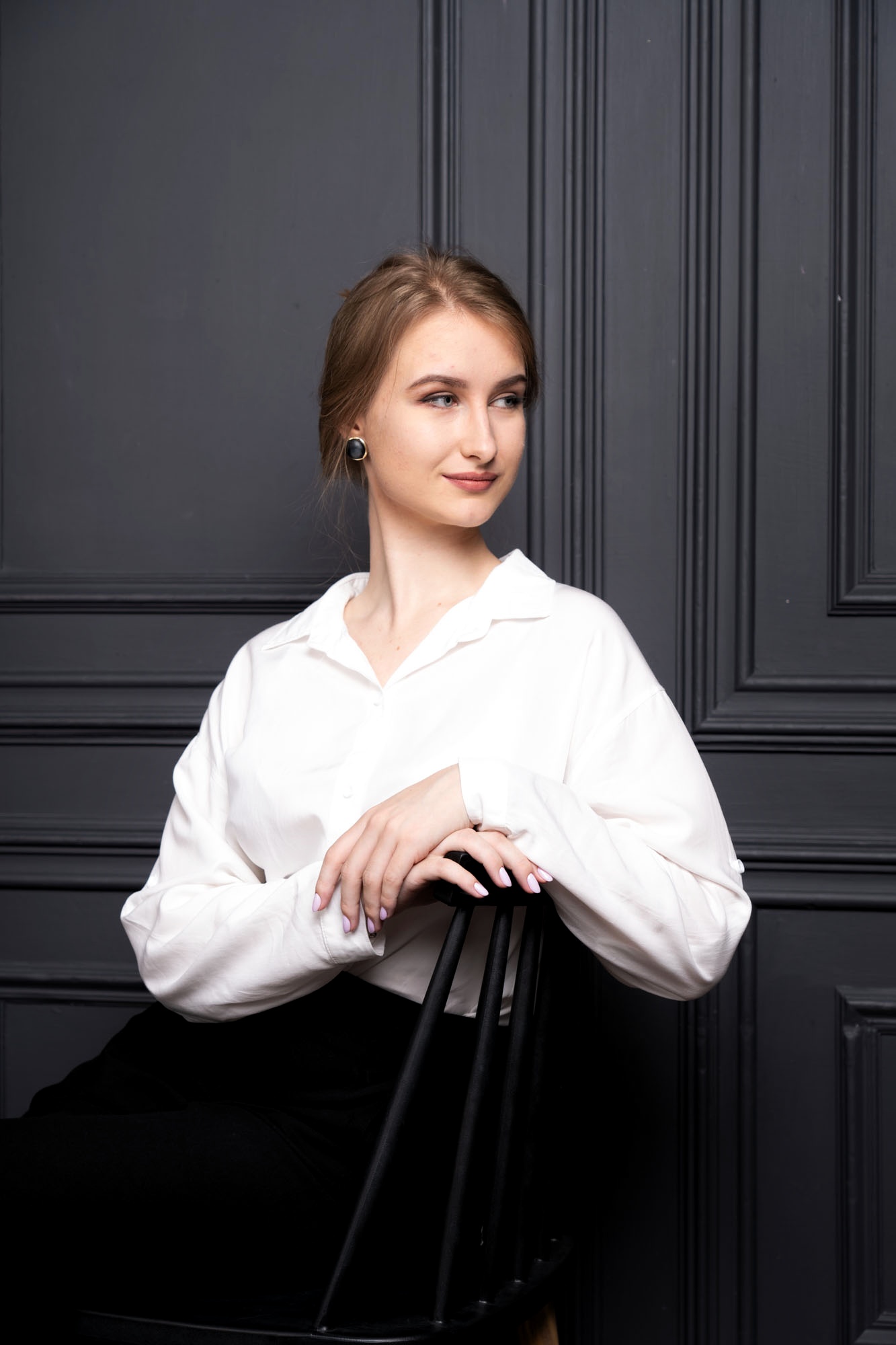Portrait of a young woman with light brown hair, wearing a white shirt, seated on a black chair against a dark paneled wall, looking to the right.