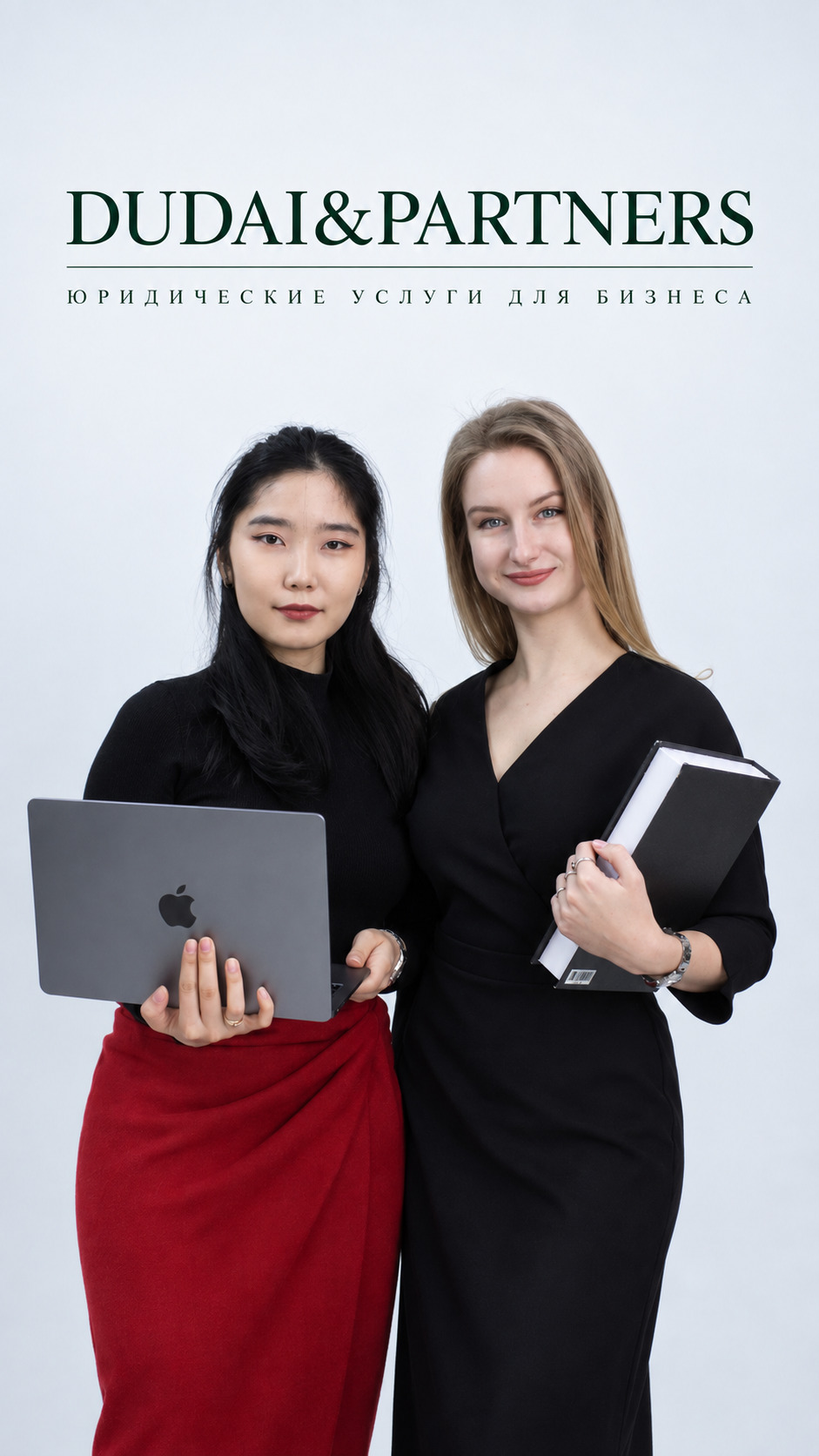 Two business professionals, a woman of Asian descent and a blonde woman, stand together holding a laptop and a notebook in front of a white backdrop with DUDAI&PARTNERS branding and Russian text beneath it.