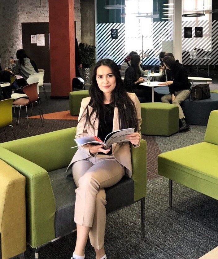 Woman in a beige suit sits on a green and yellow sofa in a modern lounge, reading a magazine.